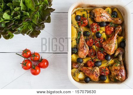 Baked chicken drumsticks in red dish. Cooked with cherry tomatoes black olives rosemary and potatoes. White wooden table top view.