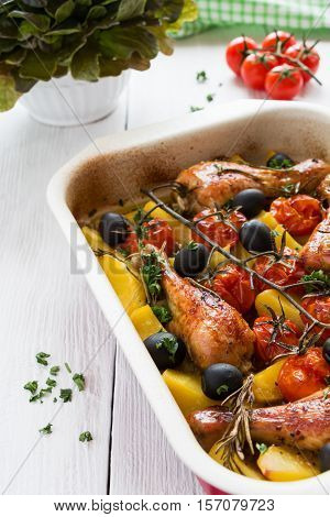 Baked chicken drumsticks in dish. Cooked with cherry tomatoes black olives rosemary and potatoes. White wooden table.