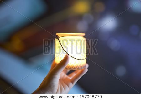 Container glass in the hand of a young woman with a candle lit inside. Horizontal shot with colors out of focus background.