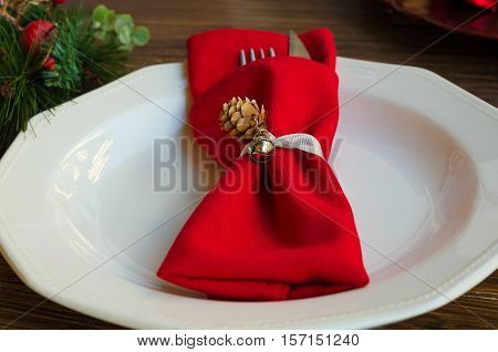 Close up of napkin ring with cone and bell on white plate. Elegant Christmas decoration for festive dinner. Red napkin with cutlery on rustic background. Copy space