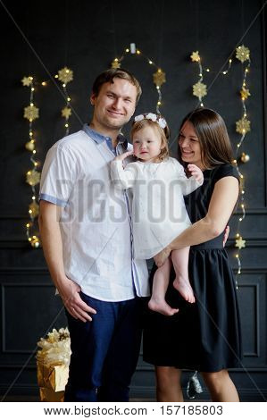 Family with little daughter is photographed in day of a holiday. Room is festively decorated. Baby is dressed in a white dress on the head of her a wreath. Girlie is capricious. Parents smile.
