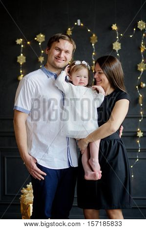 Family with little daughter is photographed in day of a holiday. Room is festively decorated. Baby is dressed in a white dress on the head of her a wreath. Girlie is capricious. Parents smile.