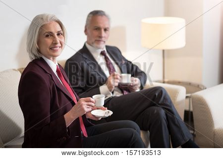 We enjoy coffee. Cheerful delighted aged businesswoman smiling and sitting in the office while drinking a coffee with her colleague and expressing happiness
