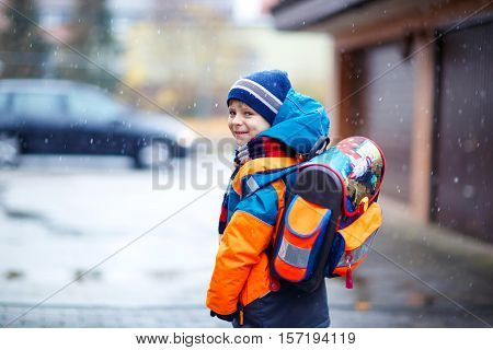 Little school kid boy of elementary class walking to school during snowfall. Happy child having fun and playing with first snow. Student with backpack in colorful winter clothes catching snowflakes.