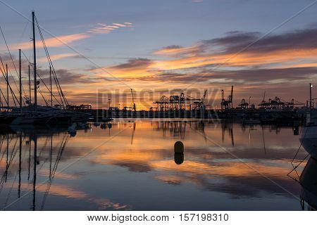 Twilight at Valencia harbor sunrises docked sailboats and cargo port cranes red colors in the sky and water reflection