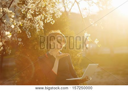 Cute girl student holding a laptop in hand. Portrait against the setting sun and cherry blossoms. She sends a kiss on the Internet. Warm toning. Videophone.