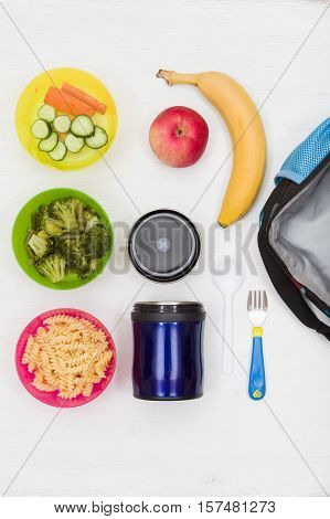 Top view on lunch box thermos and healthy food options for school. Snacks for the day. Hot lunch take away. Cucumbers carrots broccoli apple banana and pasta.