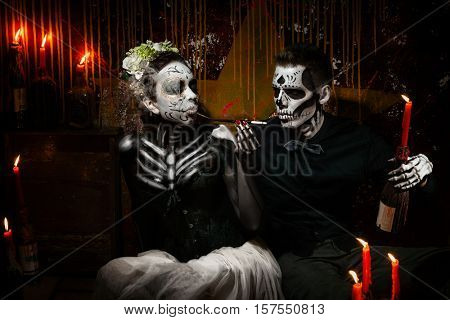 Portrait of a dead couple. Skeleton giving a light from candle to his woman with mouthpiece