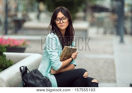 Confident asian student girl city portrait. Woman sitting on bench outside with book in casual business suite. Beautiful young mixed race Asian Caucasian woman.