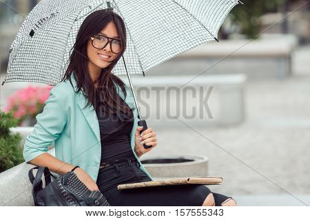 Asian girl city portrait. Woman smiling and sitting on bench outside with umbrella in casual business suite. Beautiful young mixed race Asian Caucasian woman.