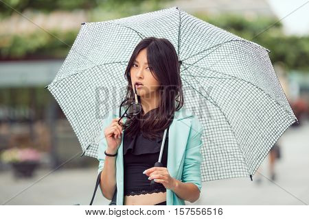 Confident asian girl city portrait. Serious sexy woman outside with umbrella and glasses in casual business suite looking at camera. Beautiful young mixed race Asian Caucasian woman.