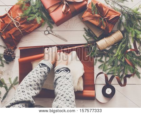 Gift wrapping background. Female hands packaging sweater as christmas present in maroon paper decorated with satin ribbon. Winter holidays concept. Top view of white wood table with fir tree branches