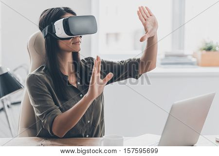 Woman in VR glasses. Confident young woman in virtual reality headset pointing in the air while sitting at her working place in office