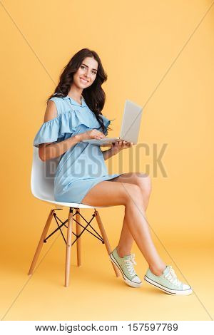 Full length portrait of a smiling brunette woman holding laptop and sitting on chair isolated on the orange background