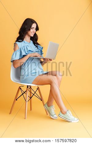 Full length portrait of a brunette smiling woman working on laptop while sitting on chair isolated on the orange background