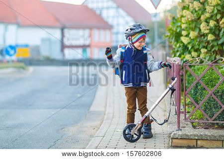 Active school kid boy in safety helmet riding with his scooter in the city. Happy child in colorful clothes biking on way to school. Safe way for kids outdoors to school.
