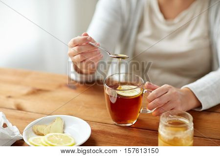 health, traditional medicine and ethnoscience concept - close up of ill woman drinking tea with lemon, honey and ginger at wooden table