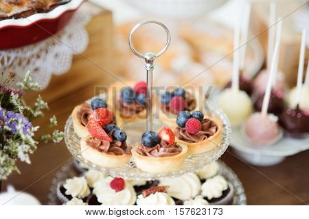 Brown wooden table with fresh fruit tarts with chocolate cream and cupcakes on cakestand. Studio shot.