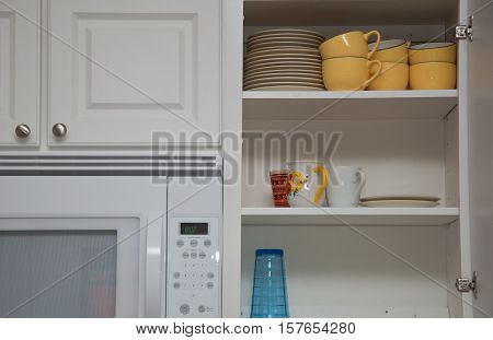 White kitchen cabinet filled with cups & plates next to a microwave