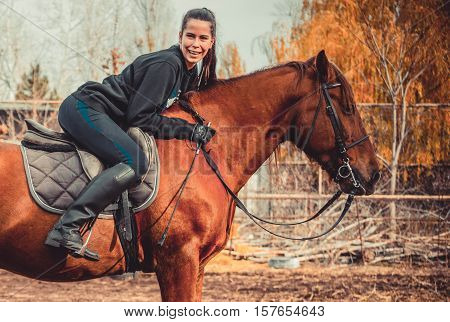 Young beautiful brunette girl rides a horse on a warm and sunny autumn day. Portrait of a pretty young woman on the horse, wearing tall boots and gloves.