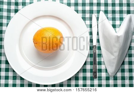 An orange on a plate with a knife & napkin on a green checkered tablecloth
