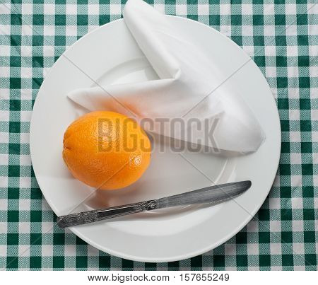 An orange on a plate with a knife & napkin on a green checkered tablecloth