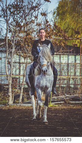 Young beautiful brunette girl rides a horse bareback with hackamore. Warm and sunny autumn day. Portrait of a pretty young woman on the horse, wearing tall boots and gloves.