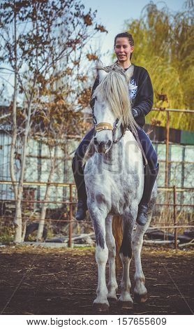 Young beautiful brunette girl rides a horse bareback with hackamore. Warm and sunny autumn day. Portrait of a pretty young woman on the horse, wearing tall boots and gloves.