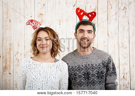 Young beautiful couple in sweaters and fake derr horns smiling looking at camera over wooden background. Copy space.