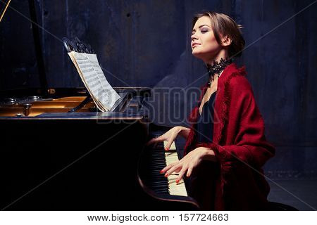 Side mid shot of delighted young woman who is making coherent sounds on the piano, enjoying the unity of melody and harmony. Wearing eveningwear with red scarf and lace black necklace 
