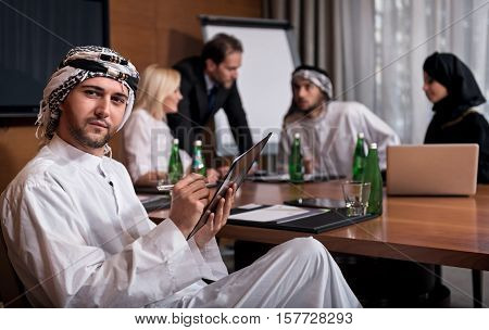 Look here. Attractive confident man participating in the meeting with four business partners wearing white robe and kaffiyeh looking forward.