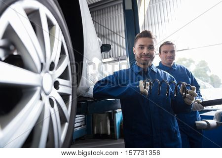 Full of positivity. Pleasant cheerful handsome mechanic holding spring and smiling while reparing car with his colleague