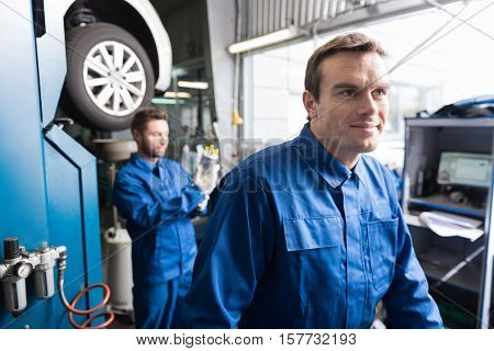 Like my work. Delighted auto mechanic smiling and standign in auto service while his colleague workign in background