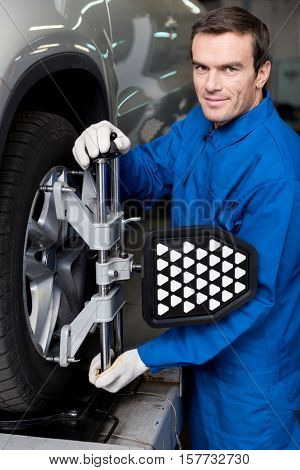 In pace with modern technologies. Pleasant adult mechanic standing near car and adjusting automobile wheel alignment while working