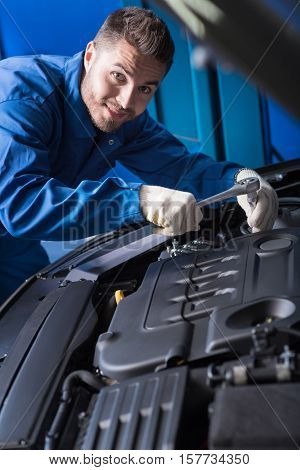 Full of gladness. Cheerful content handsome mechanic using wrench while repairing car engine