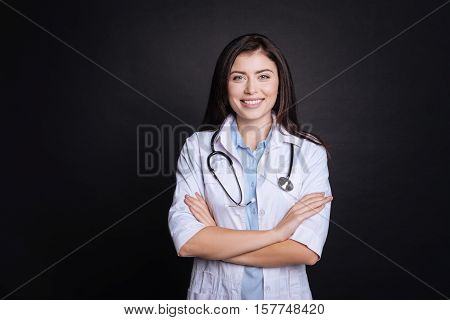 True professional.Pleasant professioanl ositive doctor folding her hands and looking at you while standing isolated on black background