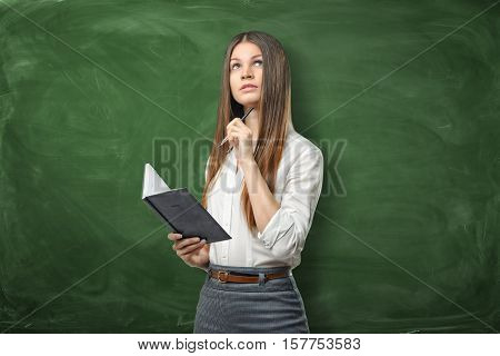 A young businesswoman holding her open day planner and a pen and going to write something on the green chalkboard background. Daily planning. Time-management. Secretary work.