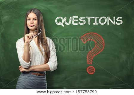 Beautiful businesswoman holding glasses in her hand and thinking about something near the green wall with big question mark written on it. Decision making. Business problems. Interviewing of respondents.
