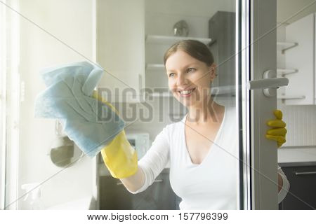 Beautiful smiling young housewife doing house chores, washing the windows, wearing rubber protective yellow gloves, with rag and spray bottle detergent. Home, housekeeping concept