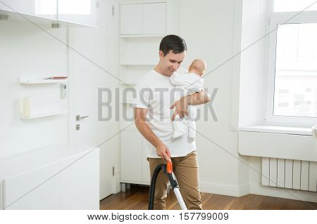 Smiling stay-at- home dad spending time together with his little child, doing vacuum cleaning carpet in living room, holding a baby in one hand and vacuum cleaner in another. Home, housekeeping concept.
