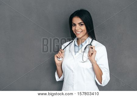 You can absolutely trust me! Attractive young female doctor in white lab coat keeping arms crossed and smiling while standing against grey background