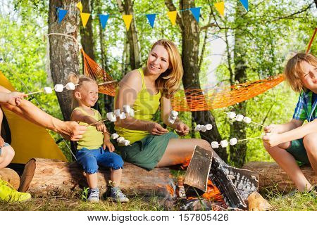 Portrait of happy young mother and kids roasting marshmallows on the sticks over the fire at campsite in summer