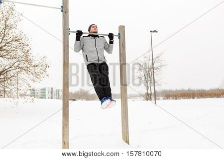 fitness, sport, exercising, training and people concept - young man doing pull ups on horizontal bar outdoors in winter