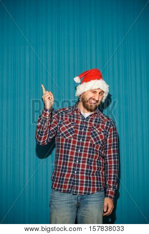 Smiling christmas man wearing a santa hat on the blue studio background pointing up
