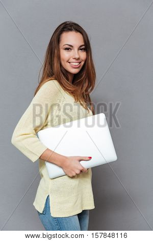 Portrait of a cheerful happy gorl in sweater holding laptop isolated on the gray background