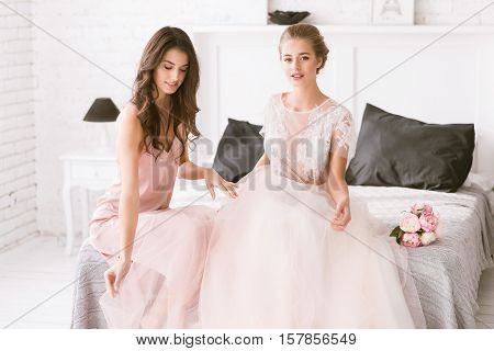 The last minutes of maidhood. Cheerful excited young bride and bridesmaid sitting in the white room while being in interaction with each other and expressing tenderness