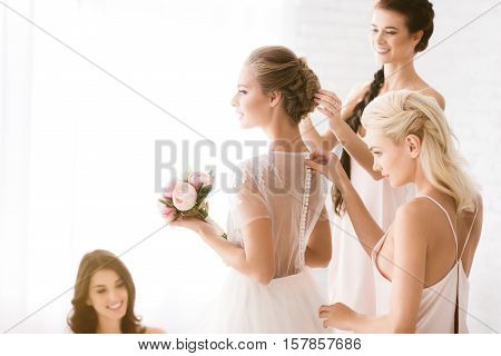 We are friends. Happy smiling young bridesmaids sitting in the white bedroom while helping the bride to get ready and expressing joy