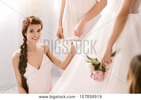 Full of happiness. Cheerful smiling young bridesmaid sitting in the white bedroom while helping with other bridesmaid the bride to get ready and expressing happiness