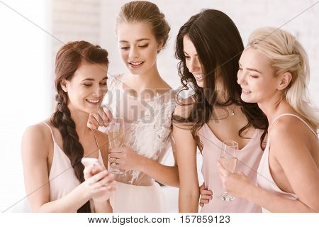 Party. Cheerful involved young girls standing in the white bedroom while looking at the phone and expressing interest