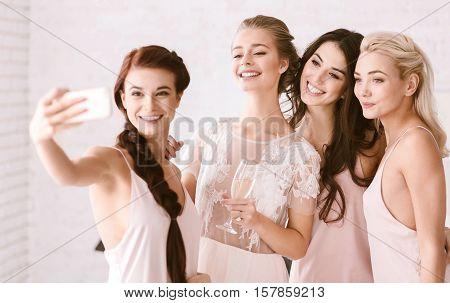 Smile. Cheerful smiling young girls standing in the white bedroom while taking a selfie and expressing joy